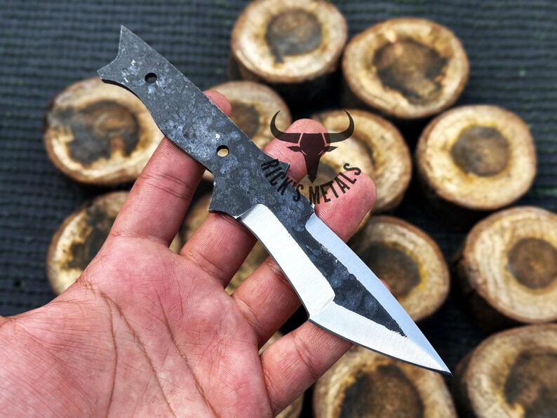 May include: A hand holds a small, black and silver knife with a sharp point. The knife has a textured handle and a partially polished blade. The background is blurred and shows a pile of wooden rounds.