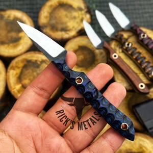 K&ouml;nnte beinhalten: Ein kleines Messer mit feststehender Klinge, einer scharfen silbernen Stahlklinge und einem strukturierten blau-schwarzen Griff mit zwei Messingnieten. Der Griff hat ein ausgepr&auml;gtes Wellenmuster. Der Text Rick's Metals ist zu sehen.