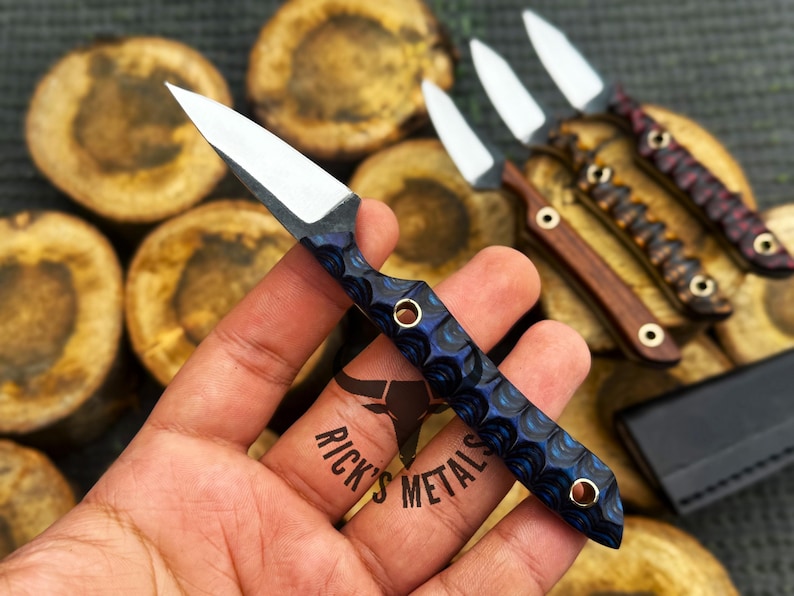 K&ouml;nnte beinhalten: Ein kleines Messer mit feststehender Klinge, einer scharfen silbernen Stahlklinge und einem strukturierten blau-schwarzen Griff mit zwei Messingnieten. Der Griff hat ein ausgepr&auml;gtes Wellenmuster. Der Text Rick's Metals ist zu sehen.