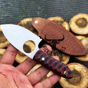 May include: A small, silver knife with a red and black handle. The knife has a finger ring on the blade and a leather sheath with a bull logo and the text "Rick's Metals".