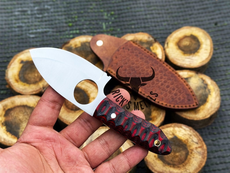May include: A small, silver knife with a red and black handle. The knife has a finger ring on the blade and a leather sheath with a bull logo and the text "Rick's Metals".