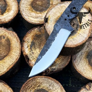 May include: A close-up of a handmade knife with a black handle and a silver blade. The knife is resting on a stack of wooden rounds. The handle has the text "Nick's Metals" on it.