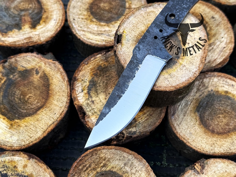 May include: A close-up of a handmade knife with a black handle and a silver blade. The knife is resting on a stack of wooden rounds. The handle has the text "Nick's Metals" on it.