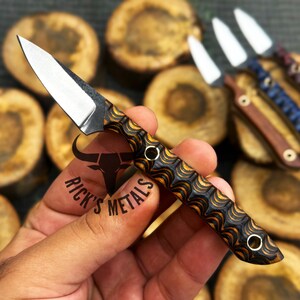 May include: A small, decorative knife with a silver blade and a handle featuring a black and gold wave pattern. The knife is held in a hand, with other knives visible in the background. The text "RICK'S METALS" is also visible.