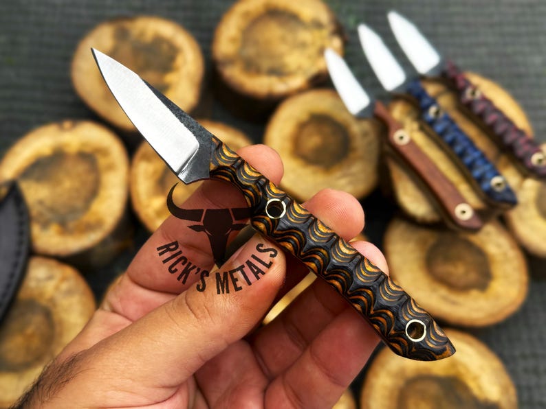 May include: A small, decorative knife with a silver blade and a handle featuring a black and gold wave pattern. The knife is held in a hand, with other knives visible in the background. The text "RICK'S METALS" is also visible.