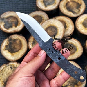 May include: A black and silver knife blade with a curved edge and a handle with two holes. The knife is held in a hand against a background of wooden rounds.
