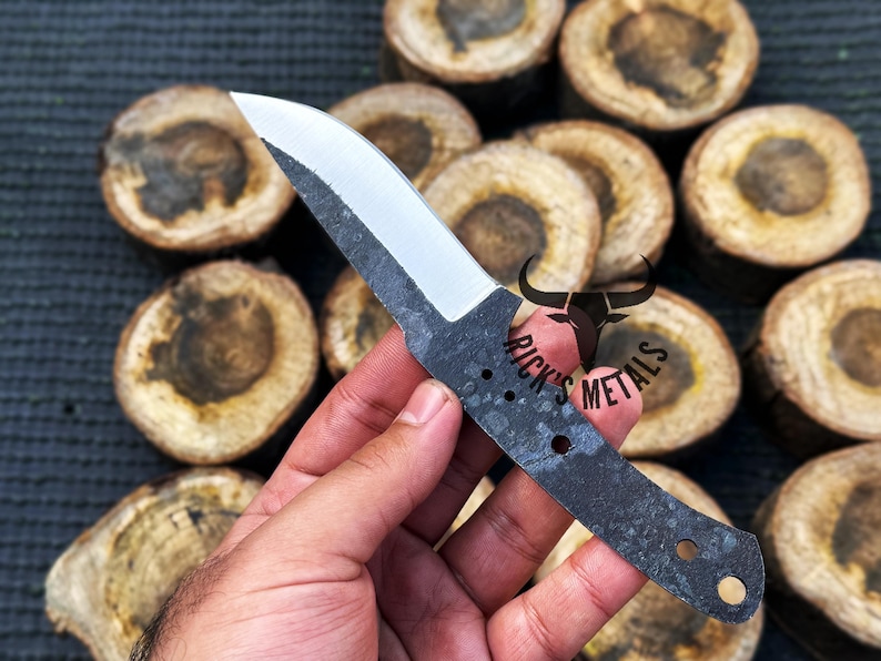 May include: A black and silver knife blade with a curved edge and a handle with two holes. The knife is held in a hand against a background of wooden rounds.