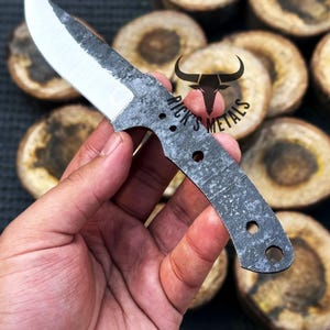 May include: A hand holds a small, gray, metal knife with a sharp blade and a textured handle. The knife has a small hole near the handle and the text "Rick's Metals" is visible on the blade.