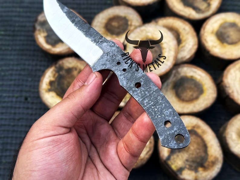 May include: A hand holds a small, gray, metal knife with a sharp blade and a textured handle. The knife has a small hole near the handle and the text "Rick's Metals" is visible on the blade.