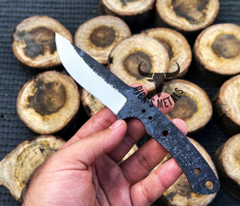 May include: A black and white knife with a textured handle and a white blade. The knife is held in a hand against a background of wooden rounds.