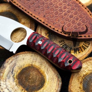 May include: A small, red and black handled knife with a finger hole in the blade. The knife is resting on a piece of wood with the text "Rick's Metals" visible.