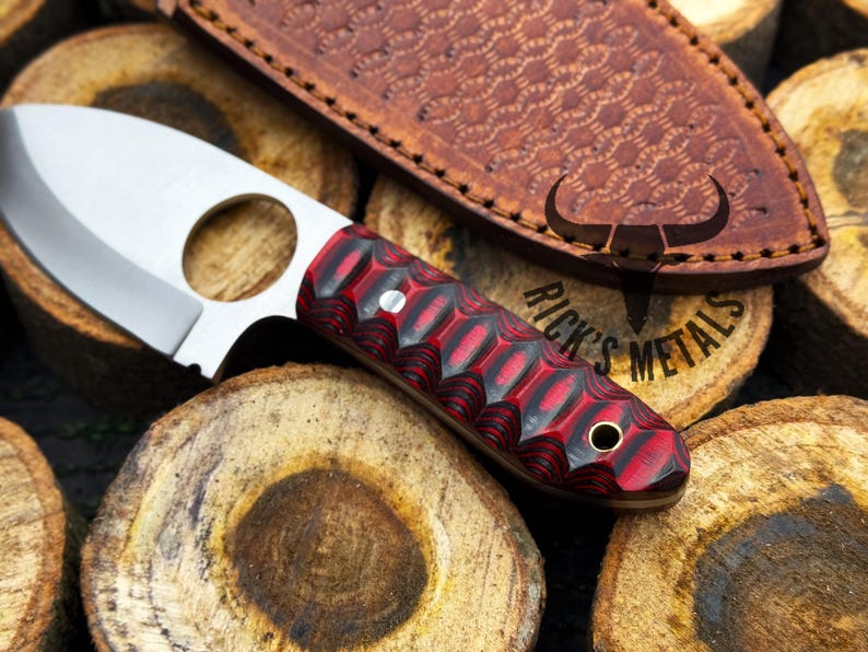 May include: A small, red and black handled knife with a finger hole in the blade. The knife is resting on a piece of wood with the text "Rick's Metals" visible.