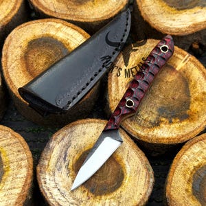 K&ouml;nnte beinhalten: Ein feststehendes Messer mit einer silberfarbenen Metallklinge und einem strukturierten, dunkelroten und schwarzen Griff mit zwei Messingnieten. Das Messer wird von einer schwarzen Lederscheide mit sichtbaren N&auml;hten und dem Logo Rack's Metals begleitet.
