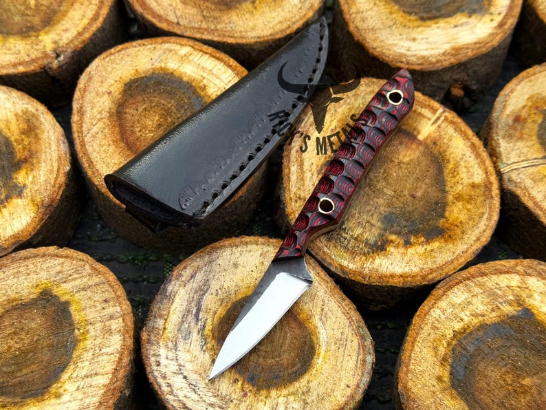 K&ouml;nnte beinhalten: Ein feststehendes Messer mit einer silberfarbenen Metallklinge und einem strukturierten, dunkelroten und schwarzen Griff mit zwei Messingnieten. Das Messer wird von einer schwarzen Lederscheide mit sichtbaren N&auml;hten und dem Logo Rack's Metals begleitet.