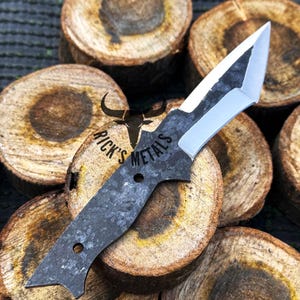 May include: A black and silver knife with a unique blade shape. The knife is resting on a stack of wooden rounds. The knife has the text "Rick's Metals" engraved on the handle.