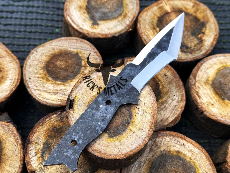May include: A black and silver knife with a unique blade shape. The knife is resting on a stack of wooden rounds. The knife has the text "Rick's Metals" engraved on the handle.