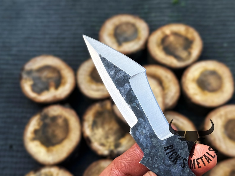 May include: A close-up of a small, black and silver knife with a unique, angular blade. The knife has a textured, hammered finish and the text "Rick's Metals" is engraved on the handle.