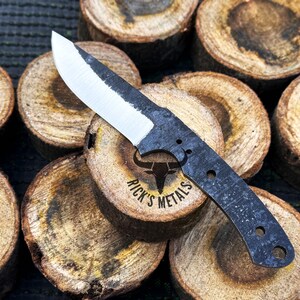 May include: A black and silver knife with a curved blade and a black handle. The knife is resting on a stack of wooden rounds. The knife has a logo that says "Rick's Metals" with a bull's head.