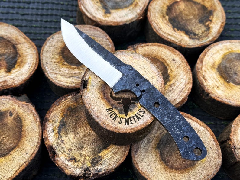 May include: A black and silver knife with a curved blade and a black handle. The knife is resting on a stack of wooden rounds. The knife has a logo that says "Rick's Metals" with a bull's head.