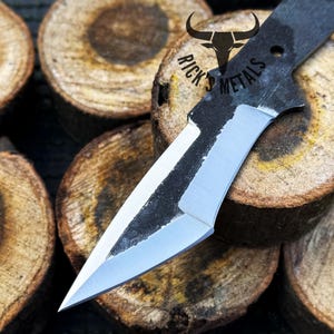 May include: A black and silver knife with a sharp point and a curved blade. The knife is resting on a stack of wooden rounds.
