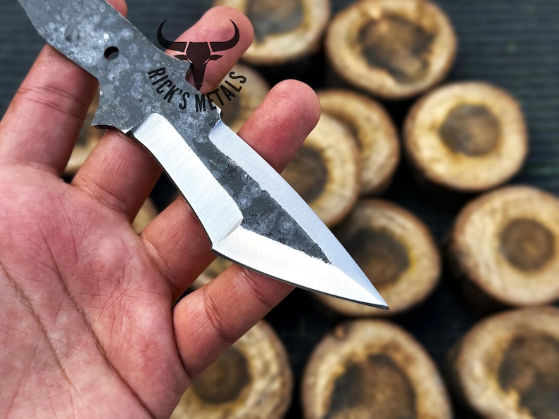 May include: A close-up of a hand holding a small, silver knife with a black handle. The knife has a sharp point and a slightly curved blade. The knife is labeled "Rick's Metals".