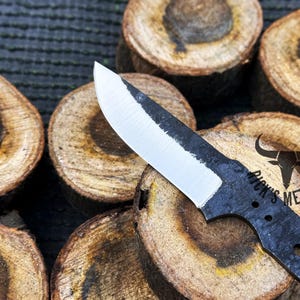 May include: A black knife blade with a white edge and a black handle. The blade has a rounded tip and a slight curve. The handle has a small hole near the end. The knife is resting on a piece of wood. The text "Dick's Metals" is printed on the handle.