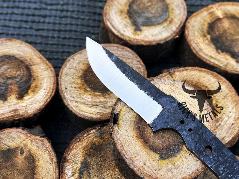 May include: A black knife blade with a white edge and a black handle. The blade has a rounded tip and a slight curve. The handle has a small hole near the end. The knife is resting on a piece of wood. The text "Dick's Metals" is printed on the handle.
