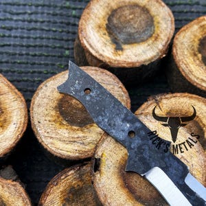 May include: A black and silver knife with a textured blade and a handle with a hole. The knife is resting on a piece of wood with the text "Rick's Metals" engraved on it.