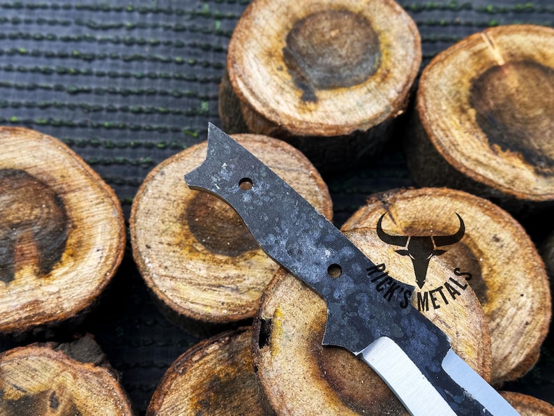 May include: A black and silver knife with a textured blade and a handle with a hole. The knife is resting on a piece of wood with the text "Rick's Metals" engraved on it.