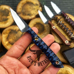 K&ouml;nnte beinhalten: Ein kleines Messer mit feststehender Klinge, polierter silberner Stahlklinge und einem strukturierten blauen Griff mit zwei Messingnieten. Der Griff hat ein gewelltes Muster. Der Text Rick's Metals ist im Hintergrund neben anderen Messern zu sehen.