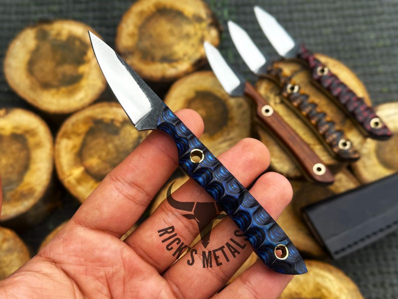 K&ouml;nnte beinhalten: Ein kleines Messer mit feststehender Klinge, polierter silberner Stahlklinge und einem strukturierten blauen Griff mit zwei Messingnieten. Der Griff hat ein gewelltes Muster. Der Text Rick's Metals ist im Hintergrund neben anderen Messern zu sehen.