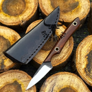 K&ouml;nnte beinhalten: Ein feststehendes Messer mit einem dunklen Holzgriff, zwei Messingnieten und einer silbernen Stahlklinge, begleitet von einer schwarzen Lederscheide mit der Pr&auml;gung RICK'S METALS und einem Stiersch&auml;del-Logo, pr&auml;sentiert auf geschnittenen Holzst&auml;mmen.