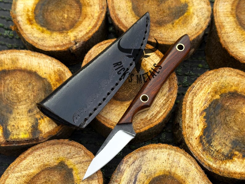 K&ouml;nnte beinhalten: Ein feststehendes Messer mit einem dunklen Holzgriff, zwei Messingnieten und einer silbernen Stahlklinge, begleitet von einer schwarzen Lederscheide mit der Pr&auml;gung RICK'S METALS und einem Stiersch&auml;del-Logo, pr&auml;sentiert auf geschnittenen Holzst&auml;mmen.