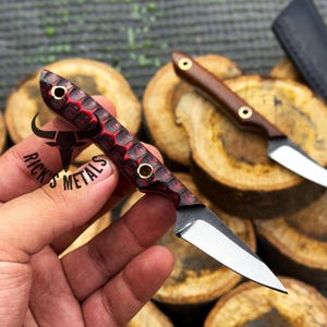 May include: A small fixed-blade knife with a textured red and black handle featuring two brass rivets. The blade is silver-colored and pointed. The handle displays the text Rick's Metals with a bull logo. A second similar knife is visible in the background.