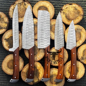 Peut inclure: Cinq couteaux de cuisine avec des manches en bois et une finition martel&eacute;e sur les lames. Les couteaux sont dispos&eacute;s sur un fond de rondins de bois empil&eacute;s. Le texte "Rick's Metals" est visible en bas de l'image.