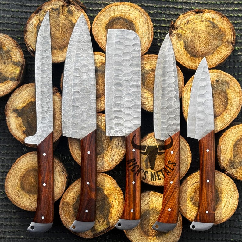 Peut inclure: Cinq couteaux de cuisine avec des manches en bois et une finition martel&eacute;e sur les lames. Les couteaux sont dispos&eacute;s sur un fond de rondins de bois empil&eacute;s. Le texte "Rick's Metals" est visible en bas de l'image.