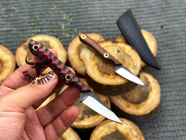 May include: Close-up of two small knives with silver blades and wooden handles. One handle is red and black, the other is brown. A black sheath is also visible. The knives are displayed on a bed of wooden slices.