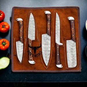 May include: A set of five kitchen knives with patterned metal blades and dark wood handles, stored in a brown leather roll-up case. The center blade features a black bull logo and the text Rick's Metals. The knives are arranged vertically.