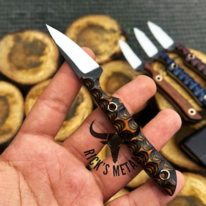 K&ouml;nnte beinhalten: Ein kleines Messer mit feststehender Klinge, einer silberfarbenen, spitzen Klinge und einem strukturierten Griff mit einem wirbelnden Muster in Schwarz und Orange. Der Griff enth&auml;lt zwei Metallnieten. Der Text RICK'S METAL ist auf dem Griff zu sehen.