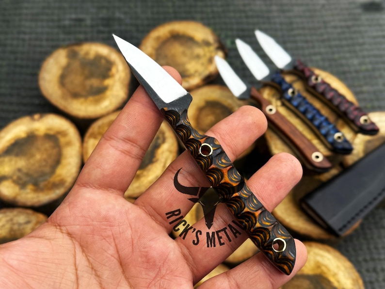 K&ouml;nnte beinhalten: Ein kleines Messer mit feststehender Klinge, einer silberfarbenen, spitzen Klinge und einem strukturierten Griff mit einem wirbelnden Muster in Schwarz und Orange. Der Griff enth&auml;lt zwei Metallnieten. Der Text RICK'S METAL ist auf dem Griff zu sehen.