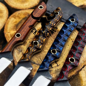 May include: Close-up of four custom knives with various handle designs. The handles feature wood grain, and patterned resin in shades of brown, blue, and red. Each knife has a silver blade and brass accents. The knives are displayed on a wooden surface.