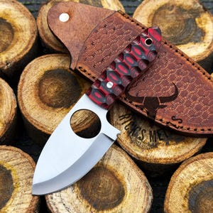 May include: A red and black handled knife with a silver blade and a leather sheath. The sheath has a bull's head design and the text "WINGS MET" is visible.