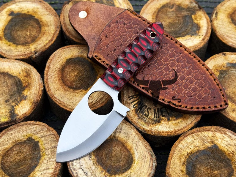 May include: A red and black handled knife with a silver blade and a leather sheath. The sheath has a bull's head design and the text "WINGS MET" is visible.