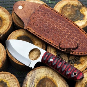 May include: A small, silver knife with a red and black handle. The knife has a finger hole in the blade and is sitting on a piece of wood. The knife is in a brown leather sheath with a honeycomb pattern.