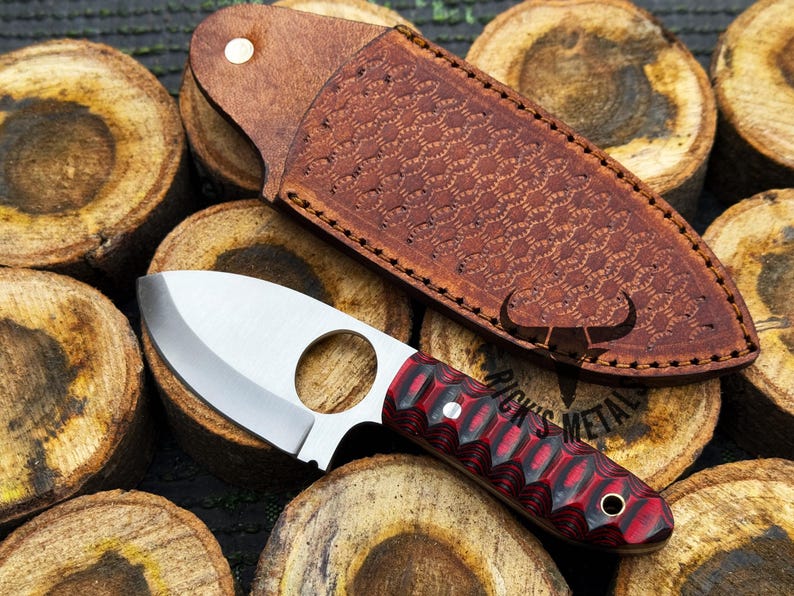 May include: A small, silver knife with a red and black handle. The knife has a finger hole in the blade and is sitting on a piece of wood. The knife is in a brown leather sheath with a honeycomb pattern.