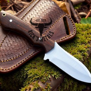 May include: A handcrafted knife with a dark wooden handle and a silver blade rests on a brown leather sheath. The sheath has a woven pattern and the text "RICK'S METALS EST. 2019" is visible on the knife.