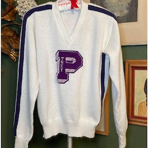 Könnte beinhalten: Weißer Vintage-Pullover mit dunkelblauen Streifen an den Ärmeln und einem violetten Filzbuchstaben 'P' mit dem Wort 'TREVY' darauf.