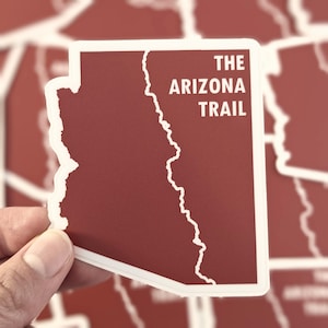May include: A sticker featuring the outline of Arizona in a deep red color, with a white border. The text "THE ARIZONA TRAIL" is printed in white. The sticker is held by a hand, with other similar stickers visible in the background.