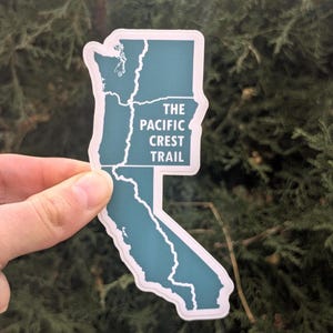 May include: A teal sticker in the shape of California, Oregon, and Washington states, with the words "THE PACIFIC CREST TRAIL" in white text. The sticker has a white border and is held against a backdrop of green foliage.