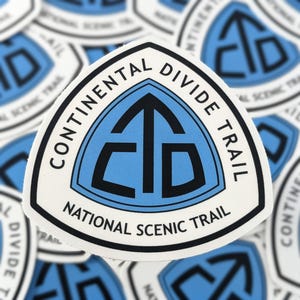 May include: A white sticker with a blue and black design. The sticker features the text "CONTINENTAL DIVIDE TRAIL" and "NATIONAL SCENIC TRAIL" in a circular pattern. The central design is a blue triangle with black lettering.